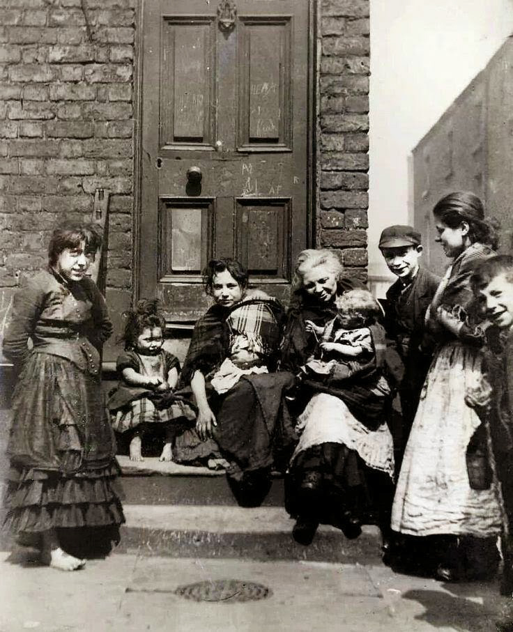 Poor women and children standing on the pavement.