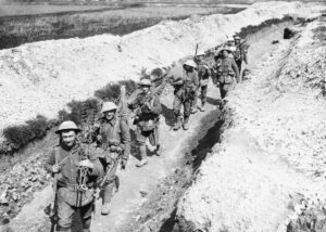 Equipped British soldiers in a trench, France, during World War I. Eight soldiers walk towards the camera in single file. They are all carrying different pieces of large equipment. The trench they are walking in is well kept and tidy. There is no mud or debris. The trench is not very deep.